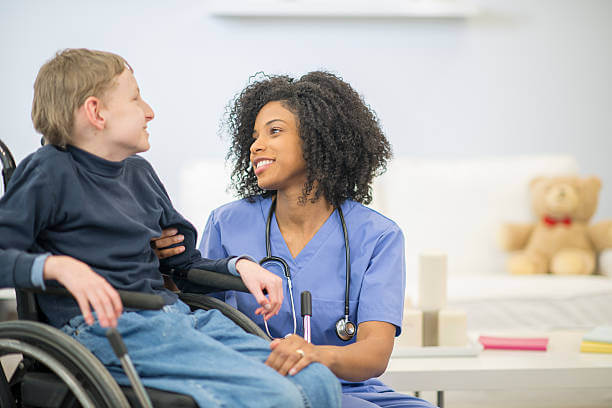 A nurse is helping a boy with cerebral palsy get into his wheelchair. They are both smiling and talking to each other.