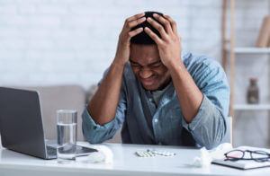 A man is experiencing a severe headache due to stress while sitting at his workplace, and he is touching his head in discomfort.